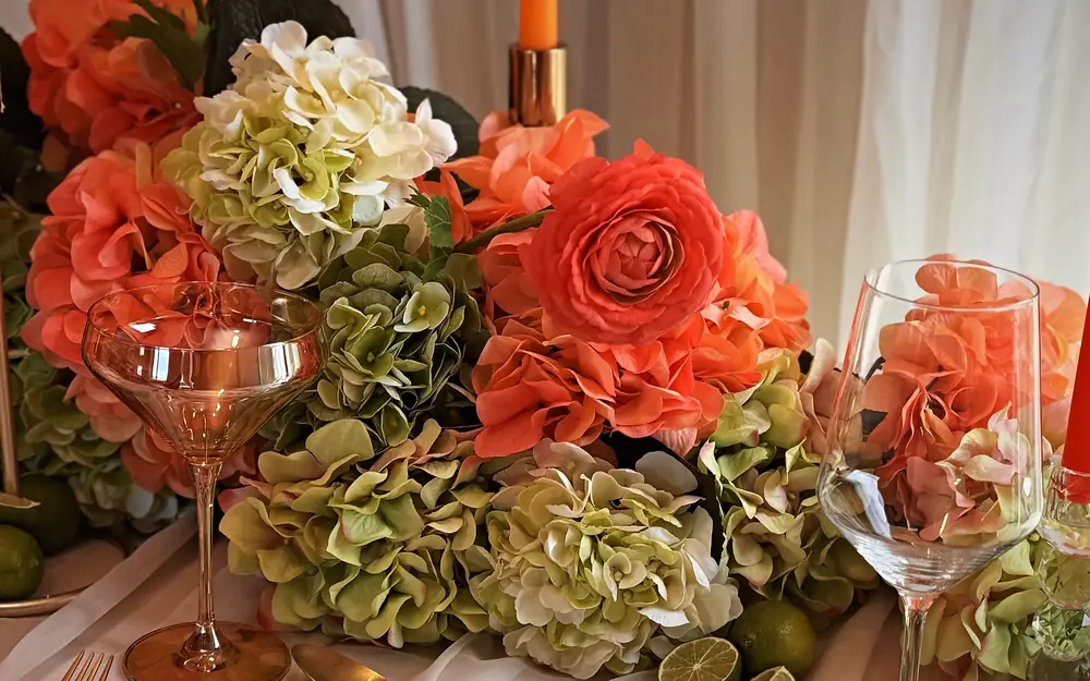 Coral ranunculus, sage hydrangeas and coupe glasses beside a lit taper on an ivory wedding tablescape, designed by Eden Events & Co.